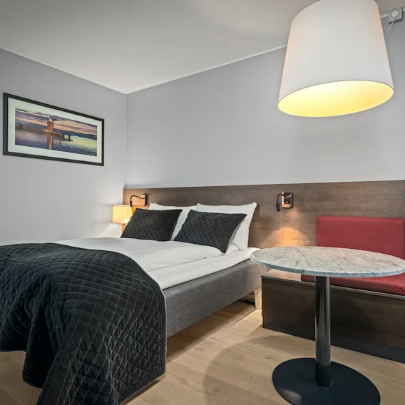 En pent oppredd seng med et mørkt teppe står mot en trehodegavel, ledsaget av et lite rundt bord og lampe i et moderne hotellrom.