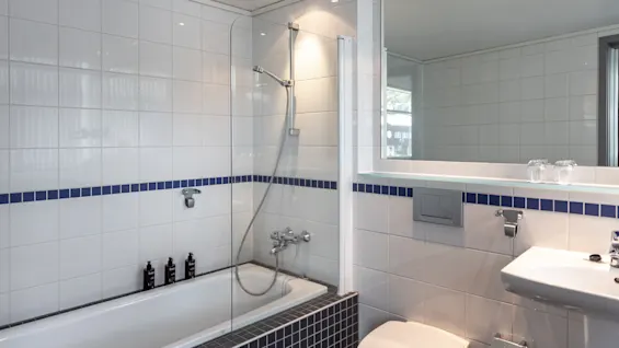 Badkar med duschhuvud, omgivet av vita plattor med blå kant. Bredvid finns en toalett och ett handfat med spegel ovanför i modern badrumsmiljö.