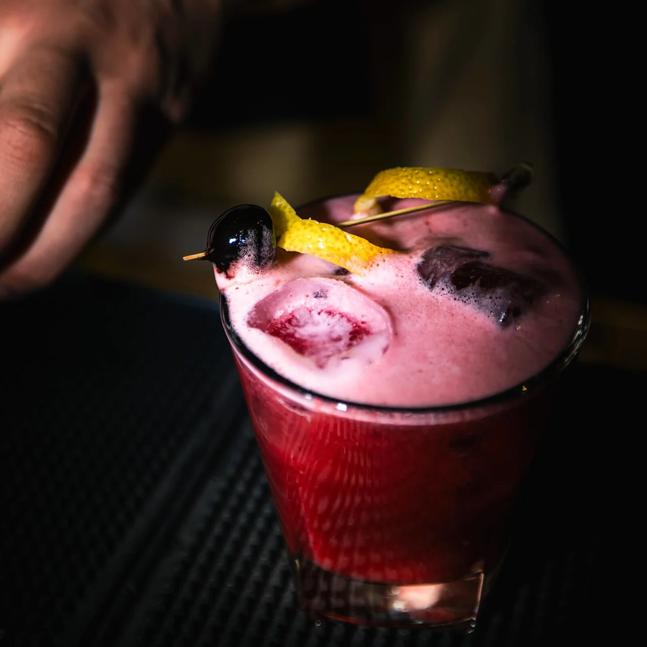 En skummande rosa cocktail i ett glas med körsbär och citrongarnityr, placerad på en svart barmatta. En hand med klocka sträcker sig mot den.