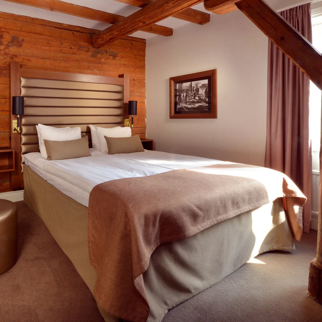 A cozy bedroom with a double bed, wooden walls, and a checkered chair, illuminated by natural light from an adjacent window.