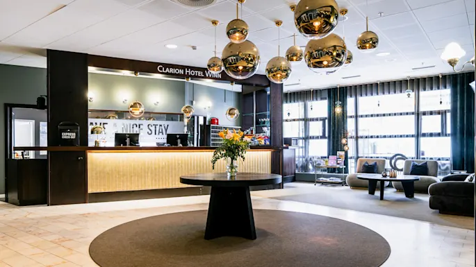 En hotellobby med et svart rundt bord og blomster, omringet av moderne sitteplasser og gyldne sfæriske lys. Skilt leser "Clarion Hotel Winn," "EXPRESS CHECK OUT," og "HAVE A NICE STAY."
