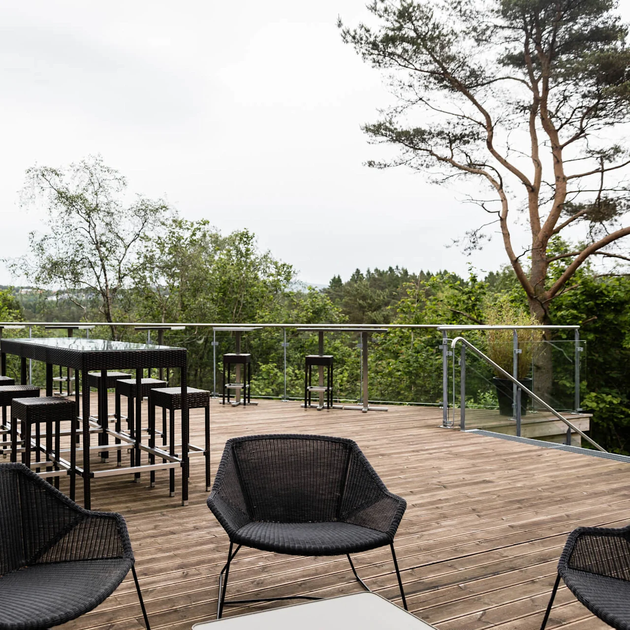 En treterrasse har svarte rottingmøbler, inkludert stoler og et høyt bord med krakker, omgitt av glassrekkverk med utsikt over et frodig, grønt skogsområde.
