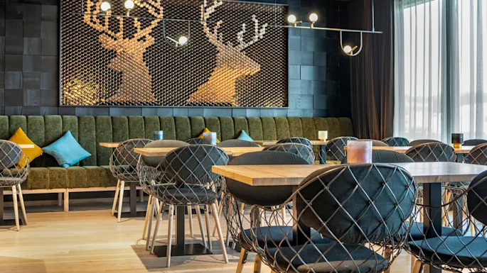 Modern dining area with wire-frame chairs around wooden tables, featuring green cushioned bench seating. Decorative wall art depicts two deer silhouettes made from circular elements, under soft lighting.