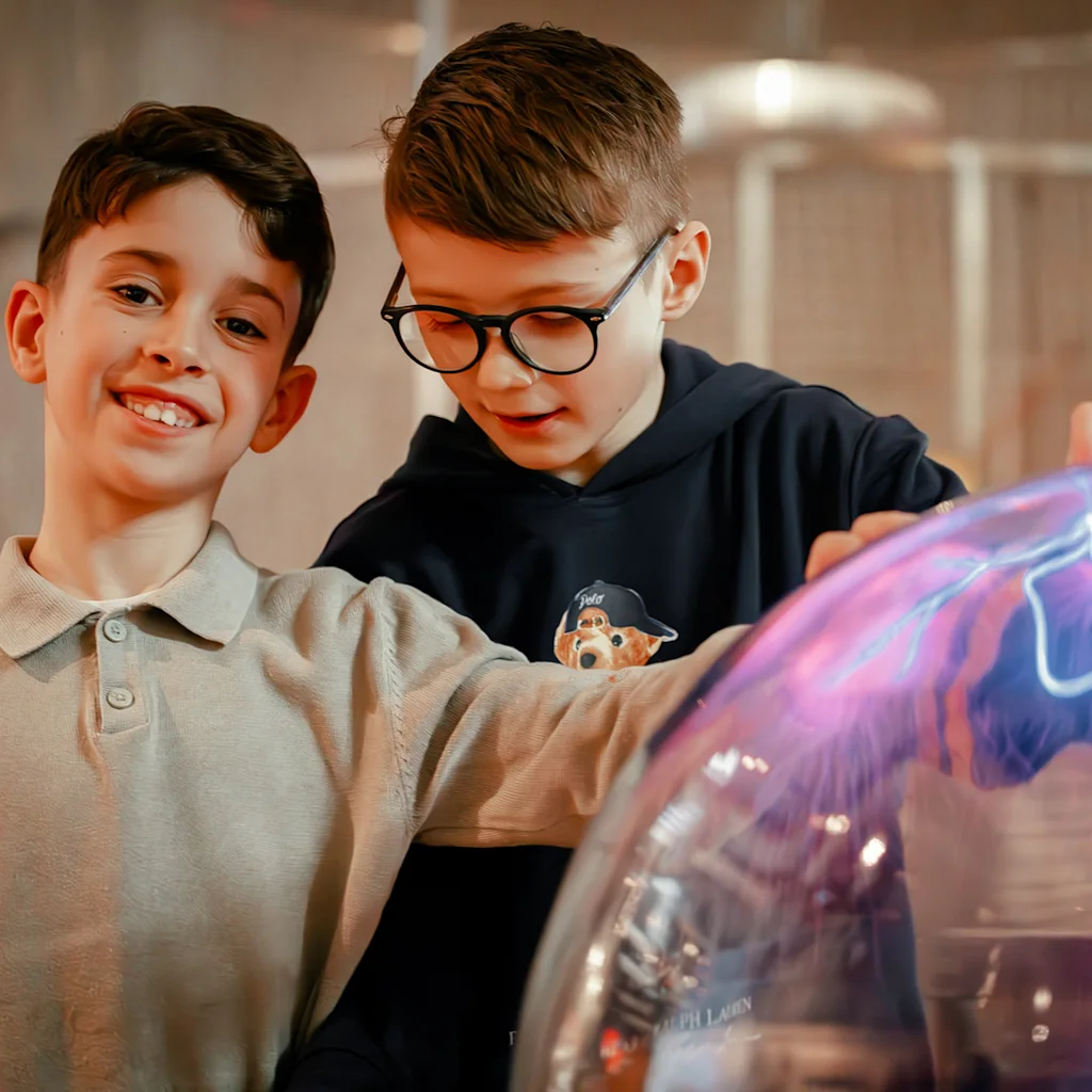 Kaksi lasta iloisesti vuorovaikutuksessa värikkään, hehkuvan tiedenäyttelyn kanssa. Toinen hymyilee kameralle, toinen on keskittynyt.