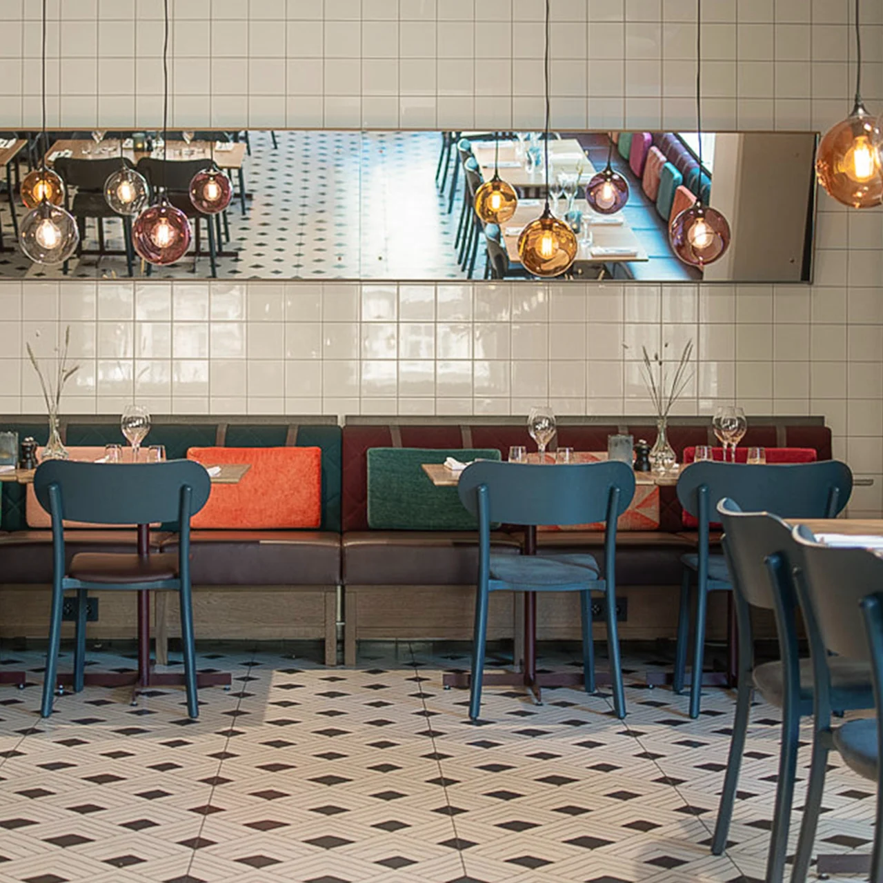 Spiseborde dækket med glas i en stilfuld restaurant; hængelamper spejles i et vægspejl. Hyndesæder og dekorative fliser forbedrer den moderne, indbydende atmosfære.