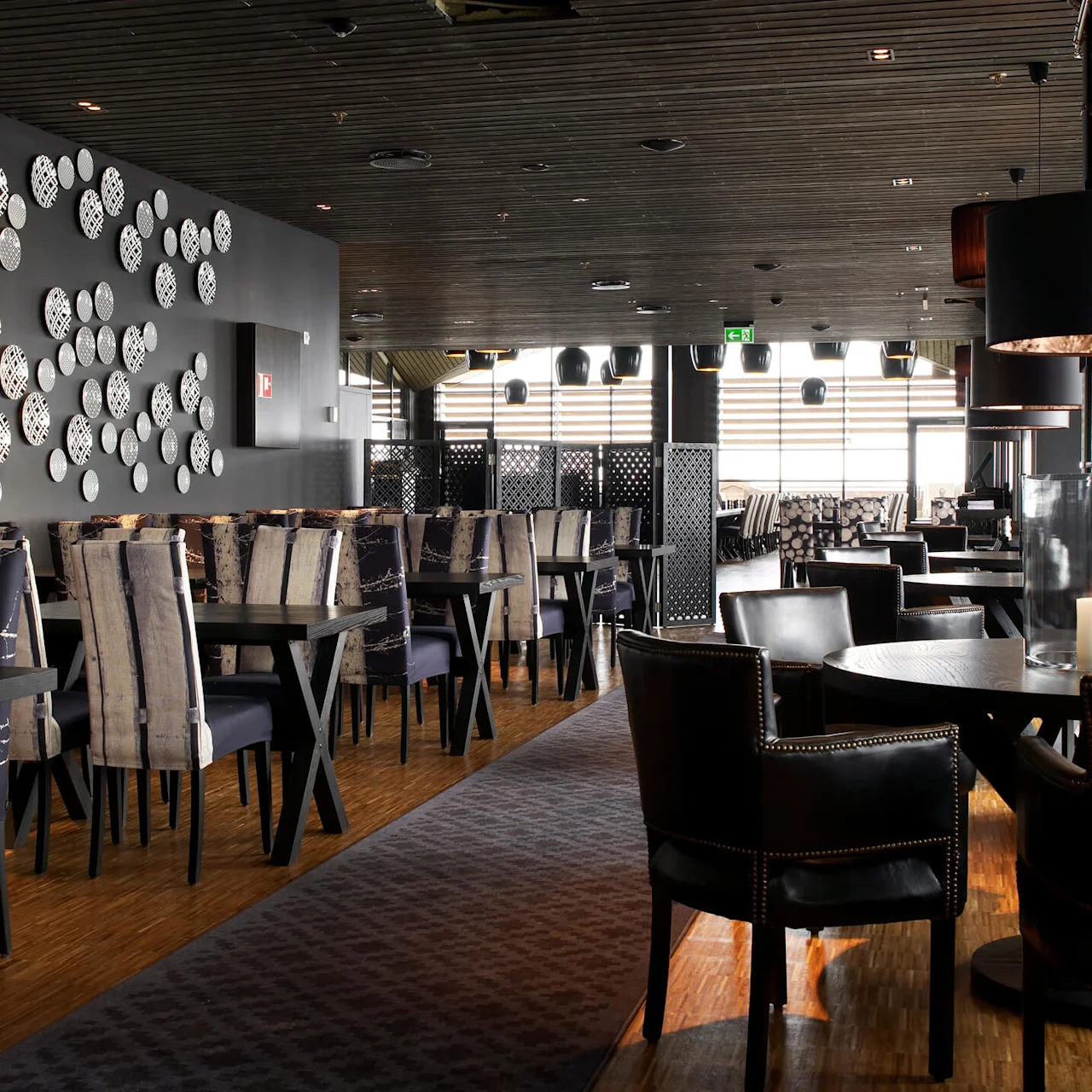 Dark wooden tables stand in a modern restaurant, adorned with patterned plates on the walls, elegant cushioned chairs, and candlelight creating a cozy atmosphere. Exit sign hangs overhead.