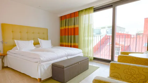 En säng med vita lakan och gul sänggavel står i ett ljust rum med stora fönster, mönstrade gardiner, fåtöljer och en grå bänk, med utsikt över röda hustak.