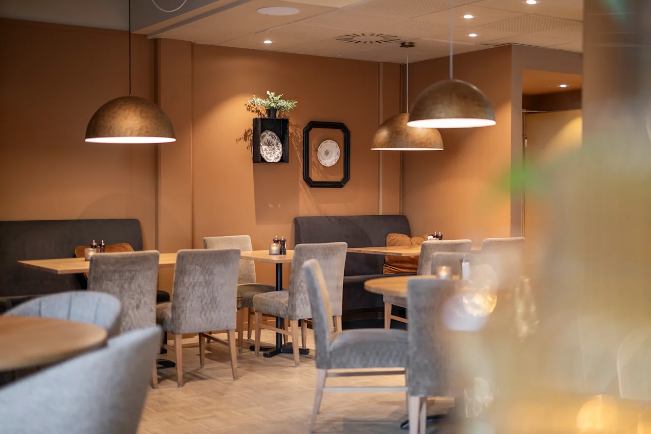 Dining tables with gray chairs are arranged in a cozy, warmly-lit restaurant. Pendant lights hang above, and decorative plates adorn the brown walls, creating an inviting atmosphere.
