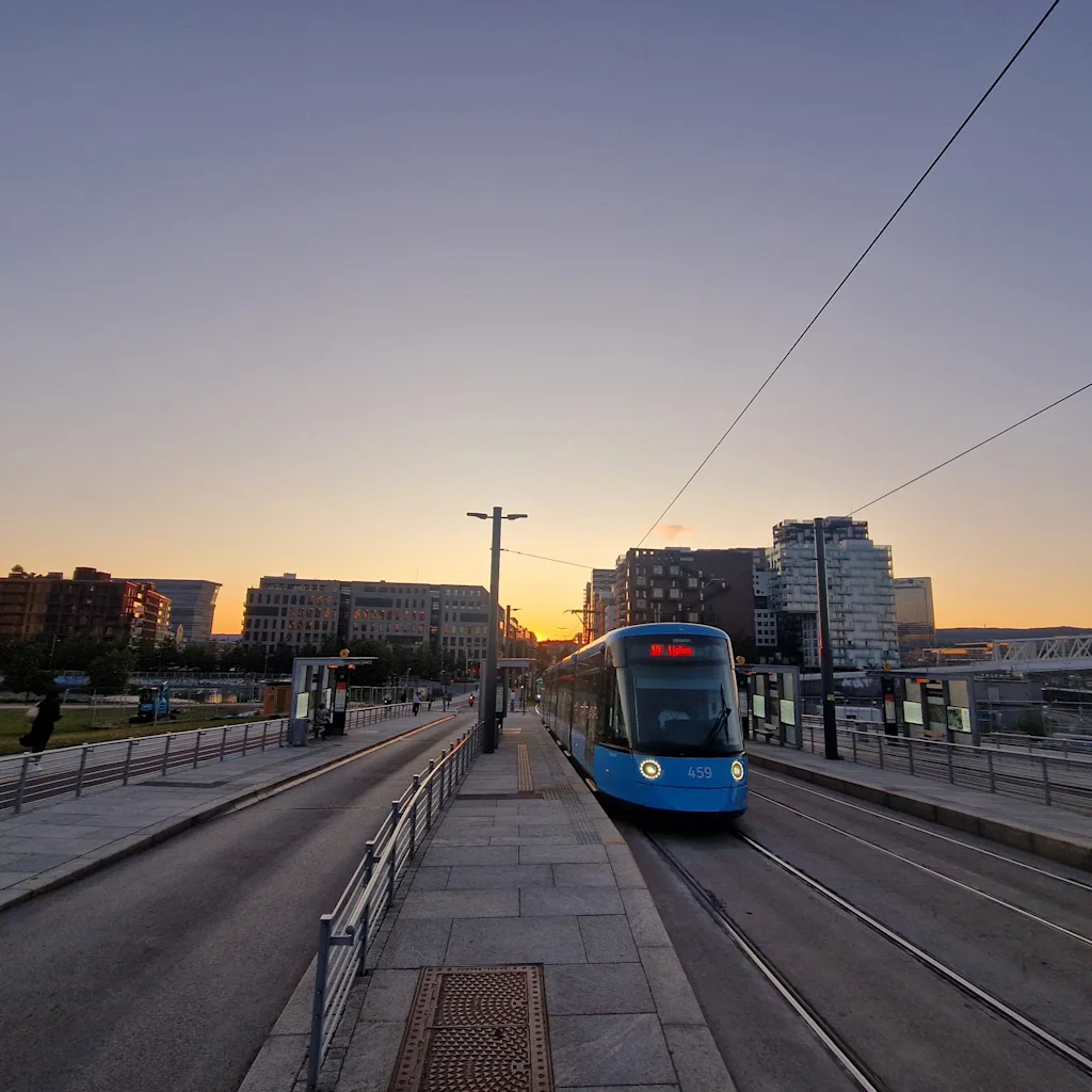 A blue tram travels on tracks through a city at sunset, with buildings and a warm sky in the background.