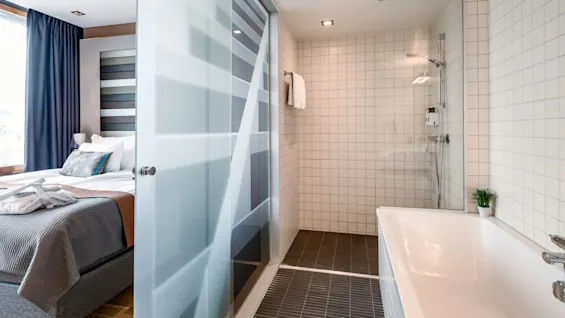 A modern bathroom features a white-tiled shower and a tub, with a glass sliding door. Adjacent, a neatly made bed with a striped headboard and large window complete the cozy bedroom.