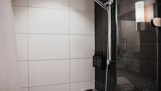 A modern shower with white and dark tiles, featuring a shower head, dispensers, and a glass screen. Home Hotel Kung Oscar.