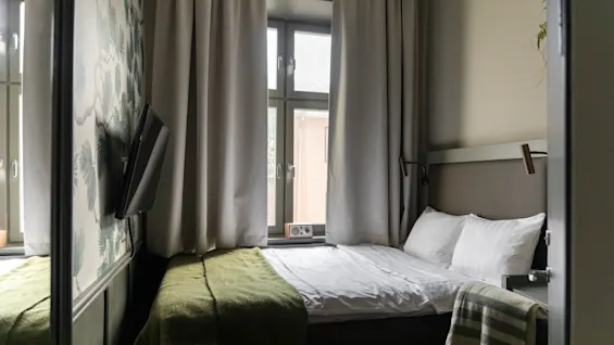 Viihtyisä hotellihuone siististi pedatulla sängyllä, ikkunalla verhoineen ja seinälle asennetulla televisiolla, luoden mukavan tilan.