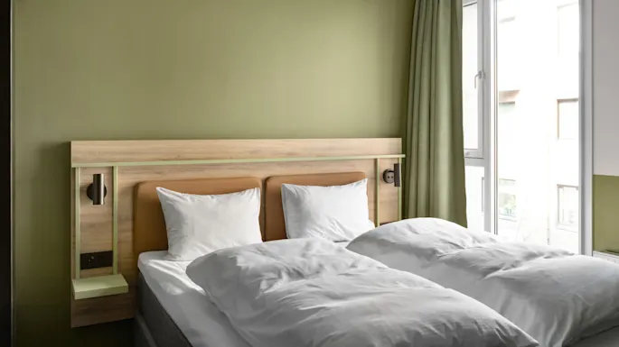Huolellisesti sijattu parivuode, jossa on valkoiset tyynyt ja peitto, seisoo vaaleanvihreää seinää vasten, lähellä ikkunaa, jossa on vihreät verhot.