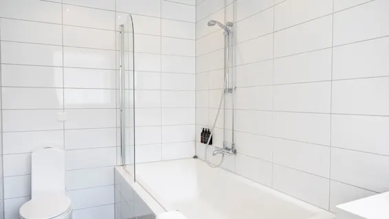Et lyst og minimalistisk badeværelse med hvide flisevægge, et badekar med bruser og et toilet, der antyder et forfriskende rum.