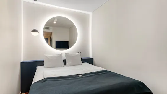 Et moderne hotelværelse med en seng, et cirkulært baggrundsbelyst spejl og en pendellampe, der skaber en hyggelig og indbydende atmosfære.