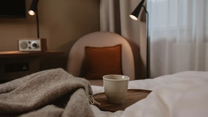 Et keramikkrus står på et trebrett på en seng med et teksturelt teppe, omgivet af en hyggelig stol, lamper og en radio i et blødt oplyst soveværelse.
