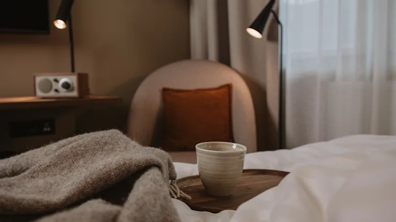 A ceramic mug sits on a wooden tray on a bed with a textured blanket, surrounded by a cozy chair, lamps, and a radio in a softly lit bedroom.