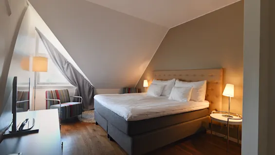 Et hyggeligt hotelværelse med en stor seng, en komfortabel stol og et skrivebord, badet i varmt lys fra lamper og et vindue.