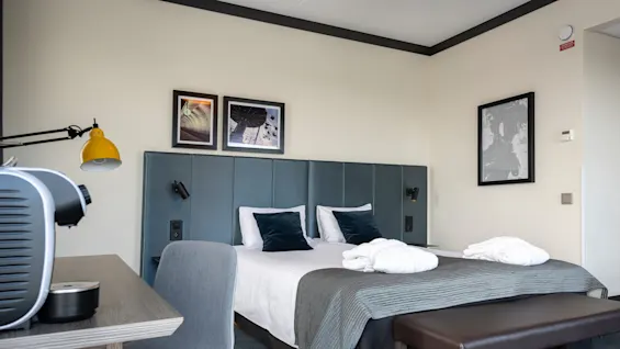A neatly arranged hotel room features a double bed with white linens and dark cushions, under framed pictures on a green headboard. Nearby, a desk holds a coffee machine.