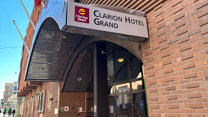 En hotellinngang med et skilt som sier "Clarion Hotel Grand." Flere flaggstenger stiger fra bygningens teglfasade i en urban setting.