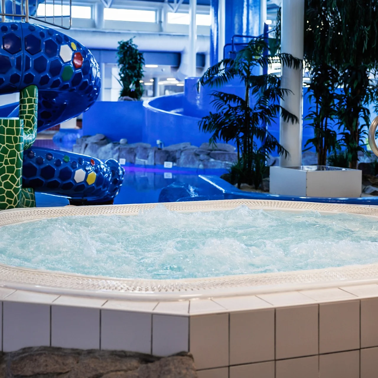 A bubbling jacuzzi sits in a bright indoor water park, surrounded by potted plants and a vibrant, twisting blue slide. 