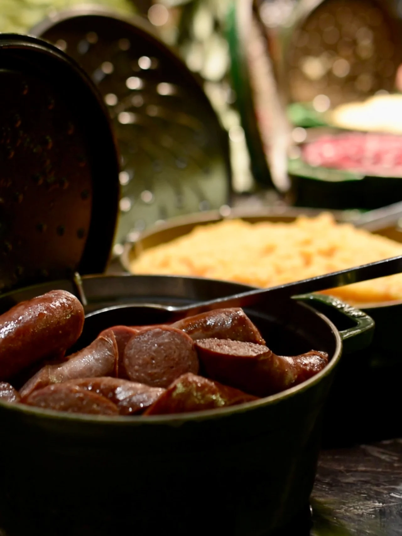 Oppskårne pølser ligger i en mørk gryte med lokk i nærheten på et buffébord omgitt av ulike fargerike retter under myk belysning.