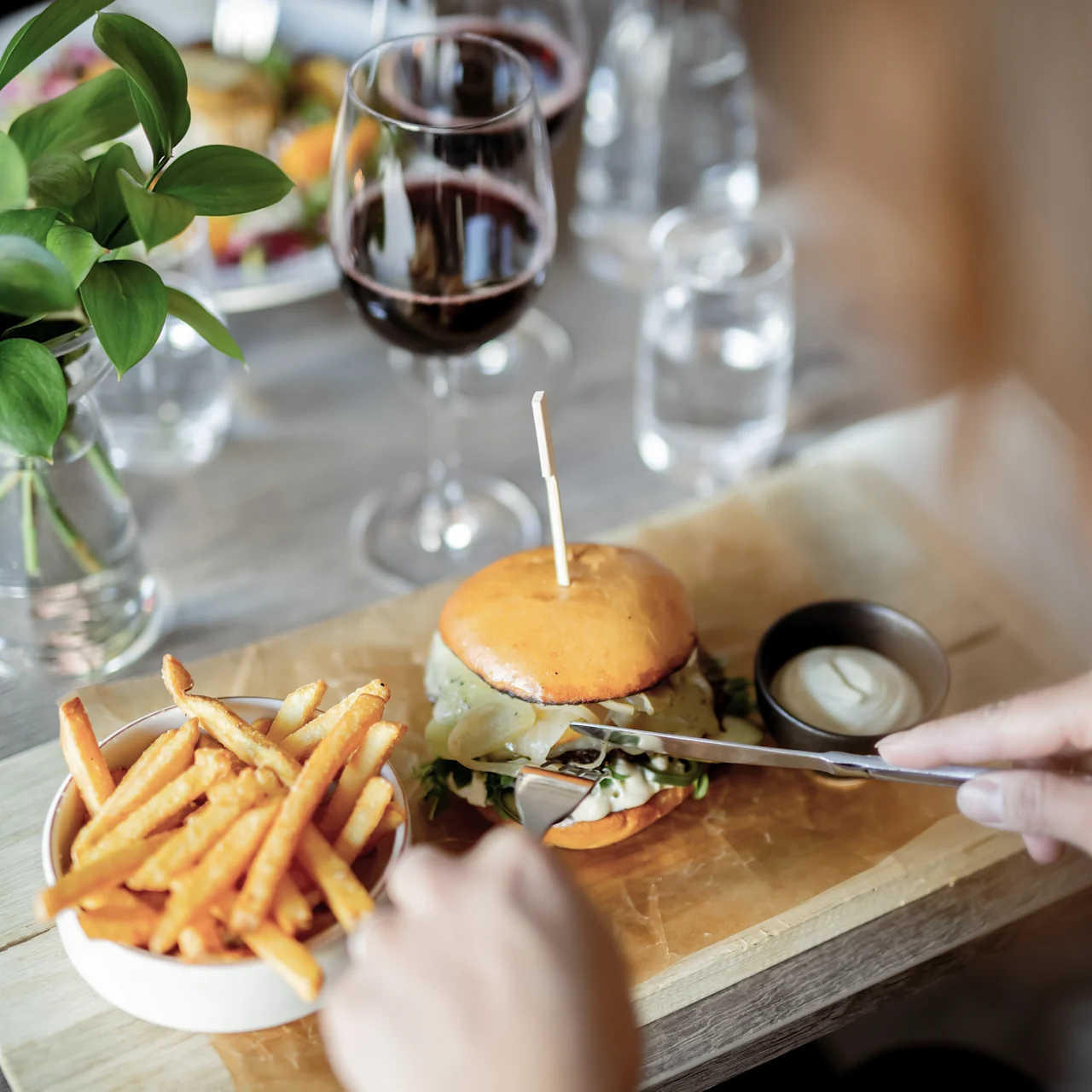 A person prepares to eat a gourmet burger and fries on a wooden board while holding a fork, with wine glasses and a plant nearby, in a restaurant setting.