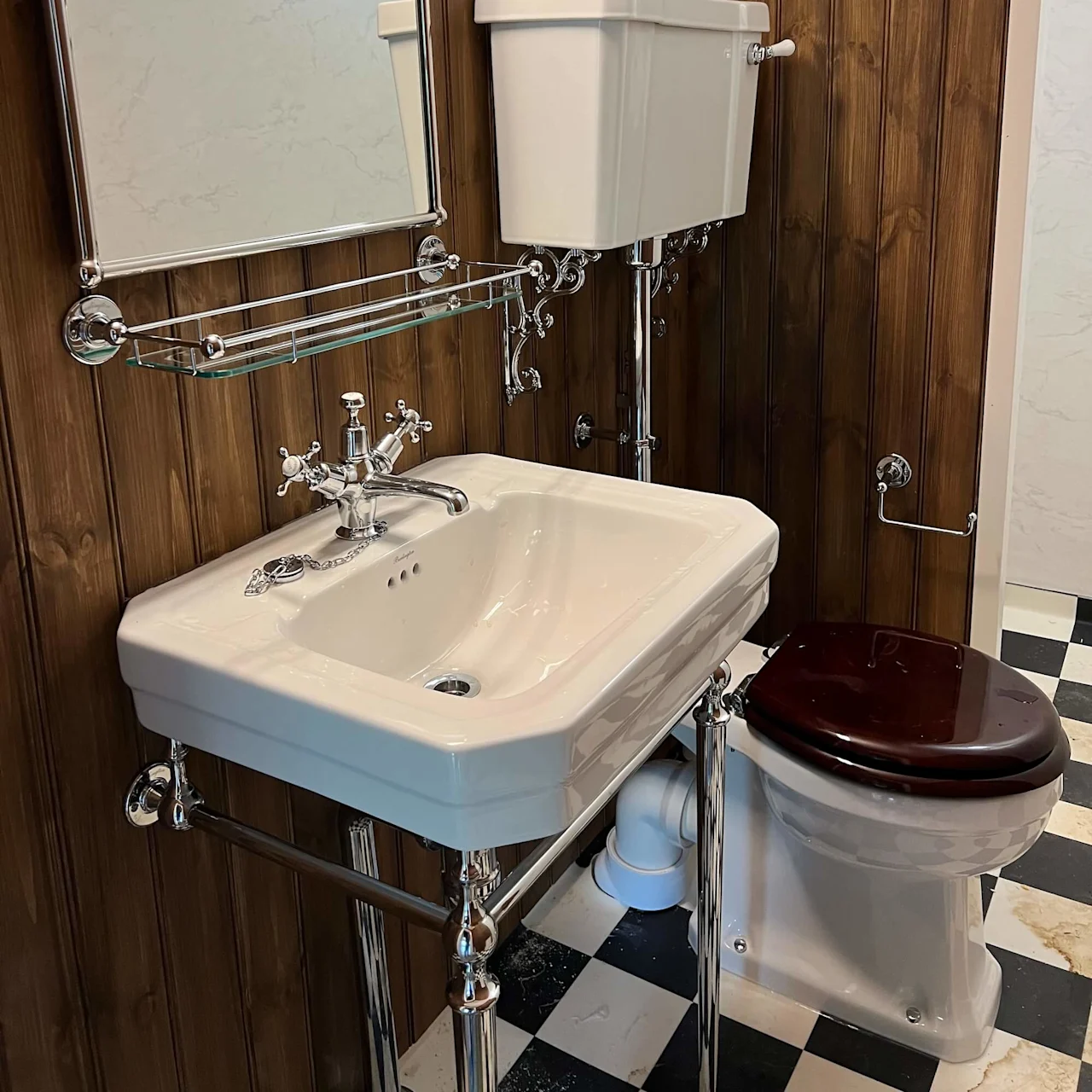 En hvid porcelænsvask med kromarmaturer er monteret på en træpanelvæg ved siden af et toilet. Et spejl hænger over, og gulvet er sort-hvidt ternet.