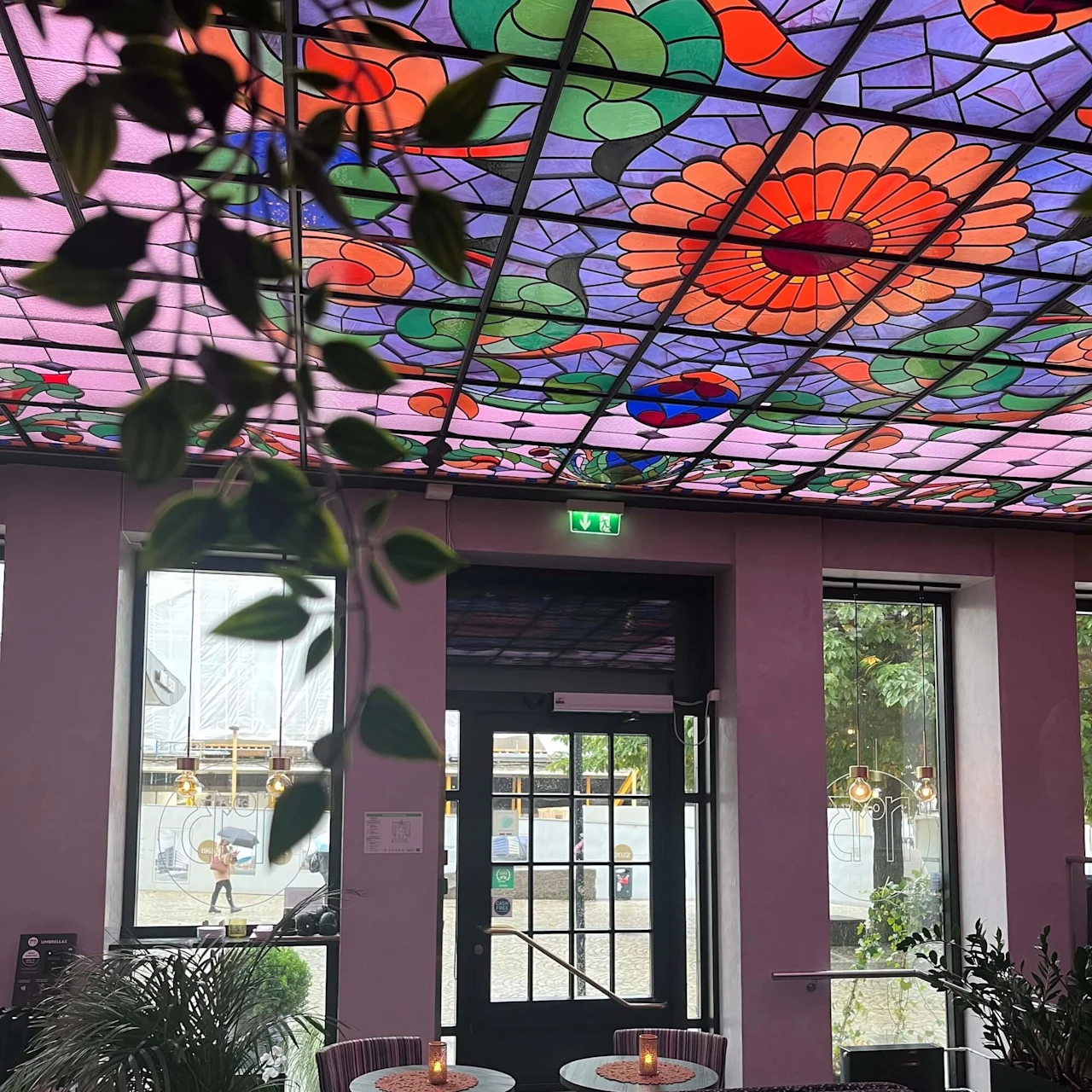 Färgat glastak med blomstermönster som lyser upp ett mysigt kafé. Ljusupplysta bord omgivna av växter och stora fönster skapar en varm atmosfär.
