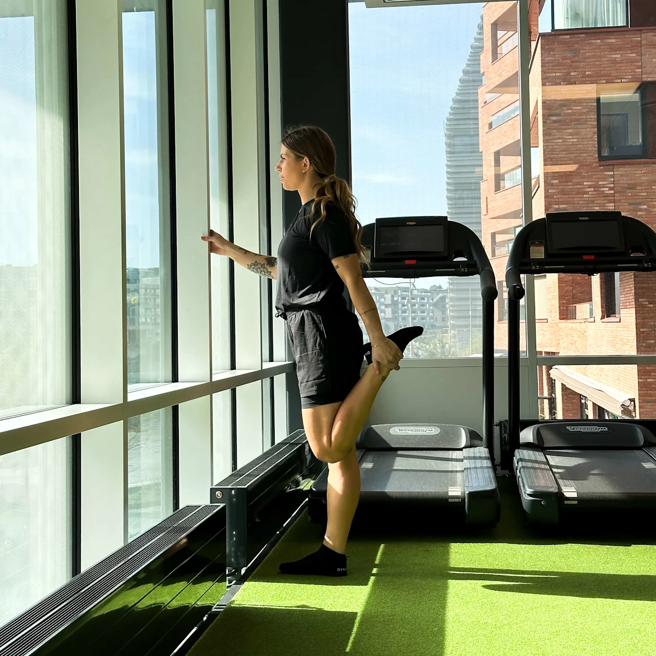 A person stretches their leg by a large window, balancing on one foot. They are in a gym environment near two treadmills, with a city view outside.