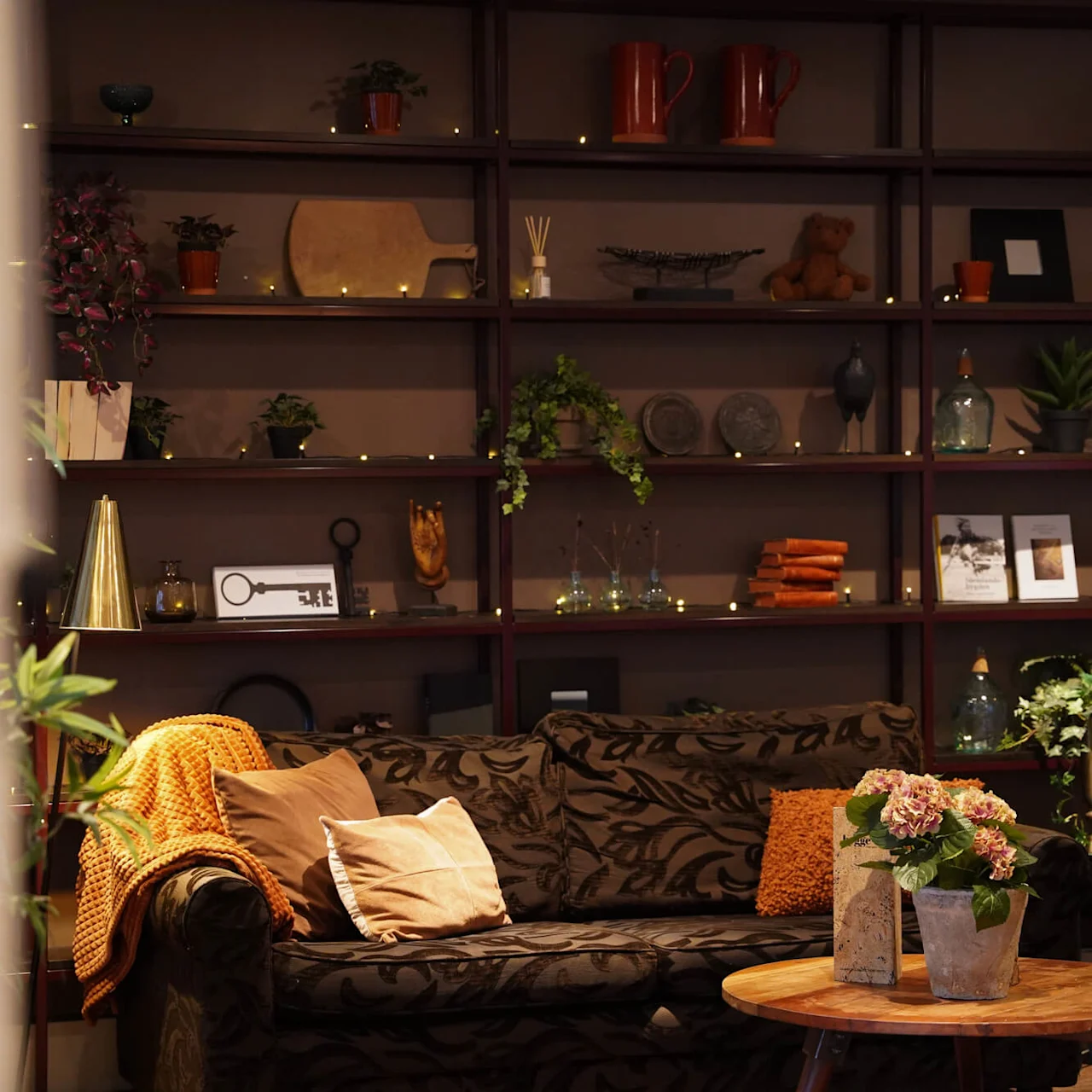 A cozy room with sofas, cushions, and a bookshelf decorated with various items and plants, suggesting a warm, homely atmosphere.