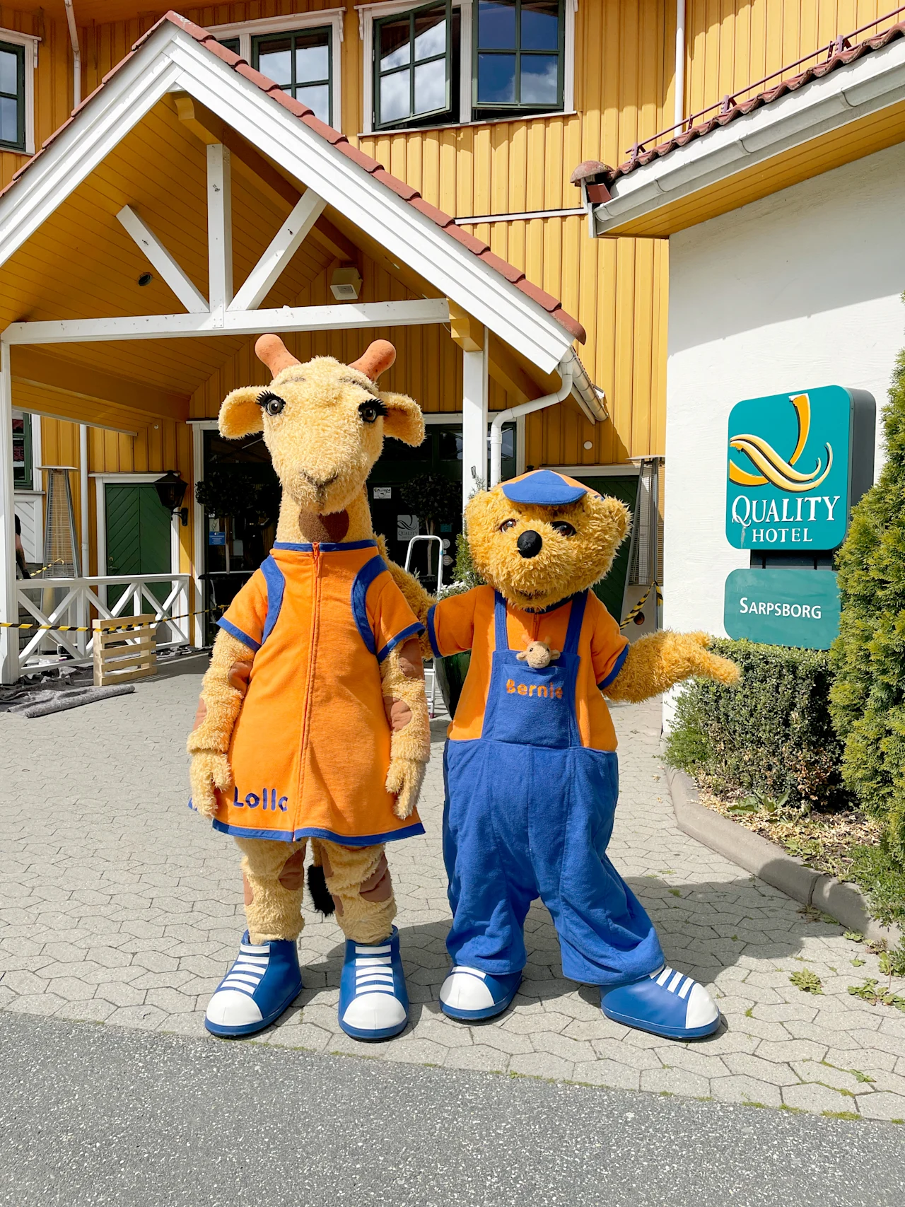 To maskoter, en sjiraff og en bjørn, står utenfor et Quality Hotel i Sarpsborg og ønsker gjester velkommen.