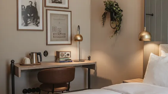 A cozy hotel room with a desk, chair, and bed. The desk has a kettle, radio, and lamp. Framed art on the wall includes "27140 344".