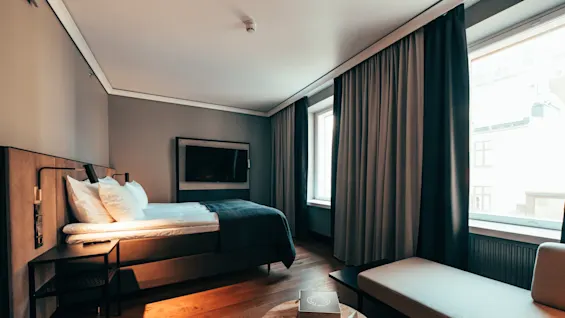 Et ryddig hotellrom med seng, bord, vegghengt TV og sofa. Naturlig lys strømmer inn gjennom to store vinduer med tykke gardiner. En brosjyre ligger på bordet.