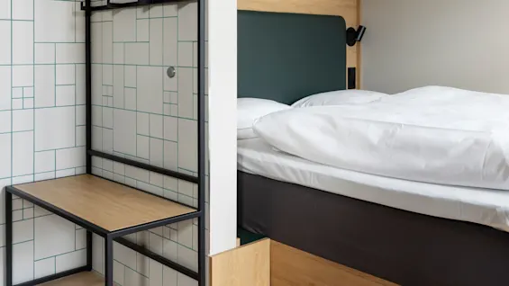 En säng med vita lakan står mot en beige vägg. Bredvid finns en svart metall- och träbänk mot en kaklad vägg med geometriska linjer.