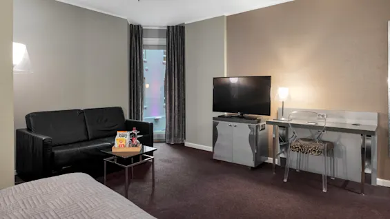 En sort sofa står i et hyggeligt hotelværelse ved siden af et lille bord med snacks. Et TV og skrivebordslampe oplyser området, suppleret af byudsigt gennem store vinduer.