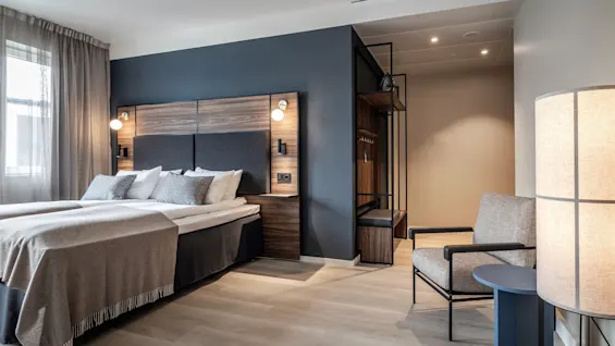 Ett modernt hotellrum med en dubbelsäng med taupefärgade sängkläder, flankerad av träpaneler och sänglampor, bredvid en sittgrupp med en stol och en golvlampa.