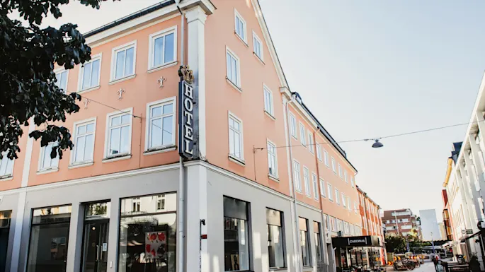 En ferskenfarget hotellbygning med et fremtredende vertikalt HOTEL-skilt på hjørnet, som ligger i en solrik gate med andre bygninger og parkerte sykler.