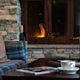 En kopp på et trebord, ved siden av et tent lys og magasiner, med en rutete sofa og steinpeis i en koselig stue.