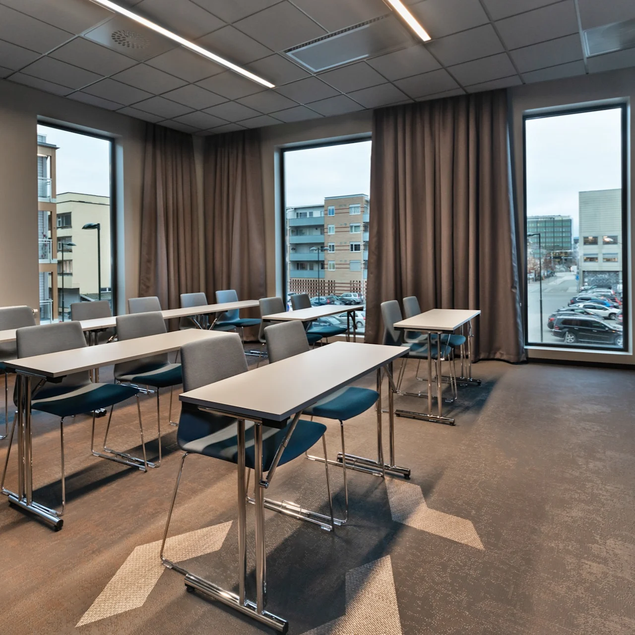 Skrivebord og stoler er ryddig arrangert i et klasserom, med store vinduer som viser en byutsikt med parkerte biler og omkringliggende bygninger. Ingen synlig tekst.