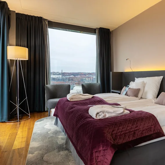 Two neatly arranged beds with burgundy quilts and pillows, situated in a modern room with a large window showing a cityscape. A floor lamp and armchair are nearby.