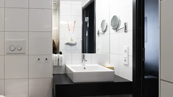 En vit handfat med kromkran är monterad på en svart bänkskiva i ett kaklat badrum. Väggmonterade flaskor är märkta "Shampoo" och "Conditioner", och det finns runda speglar.