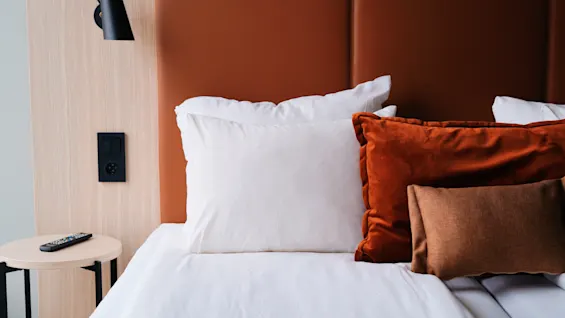 Et koselig hotellrom på Quality Hotel Arlanda XPO med en seng, hvite og oransje puter, og en fjernkontroll på et nattbord.