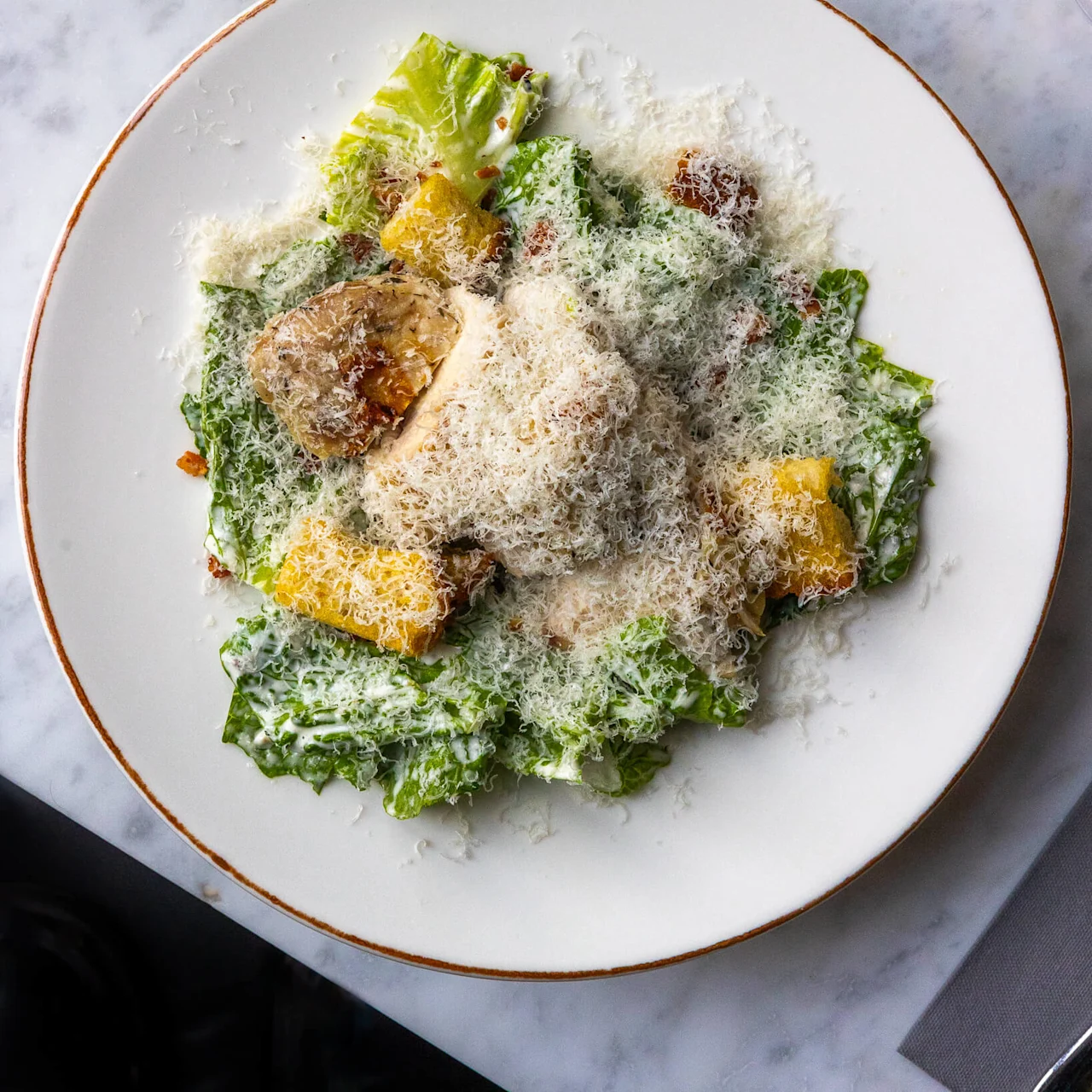 A Caesar salad topped with grated cheese and croutons sits on a white plate. The dish is placed on a marble surface, flanked by empty wine glasses.