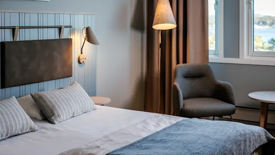 A bed with striped pillows and a blue-gray blanket positioned beside a reading chair and lamp in a cozy, softly lit room with light walls, curtains, and a window view.