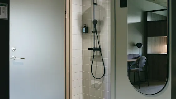 A modern bathroom with an open door revealing a tiled shower area with a black shower head and a mirror reflecting a room.