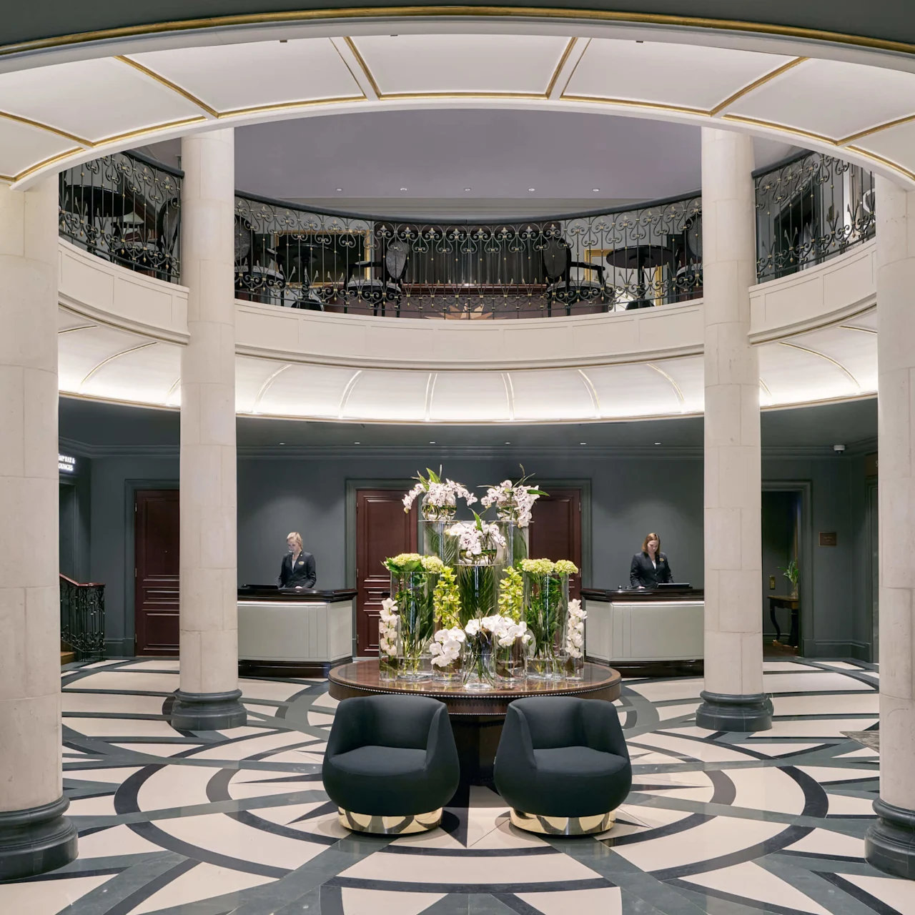 Elegant hotel lobby with two reception desks, a central floral arrangement, columns, and stylish seating, under a mezzanine balcony at Hotel Kämp in Helsinki.