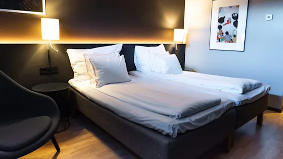 En prydligt bäddad dubbelsäng med vita kuddar och lakan står mellan två tändade vägglampor i en modern sovrumsmiljö, med abstrakt konst på väggen till höger.