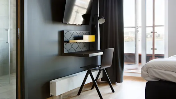 En väggmonterad TV vetter mot ett modernt skrivbord med stol och lampa nedanför, i ett rum med balkongutsikt genom en glasdörr.