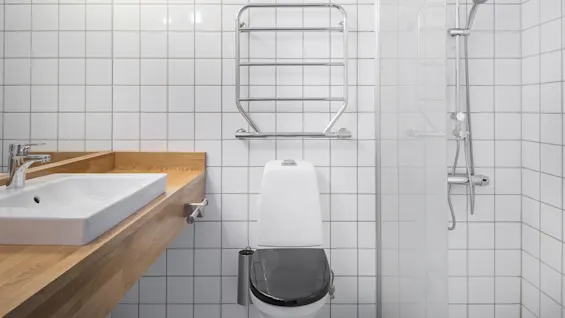 Toilet positioned against white tiled wall, beneath a metal towel rack; adjacent are a wooden sink counter and glass shower, creating a modern bathroom interior.